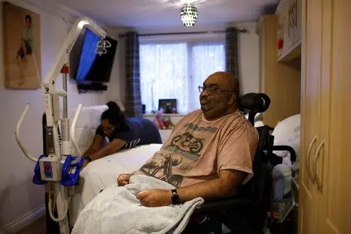 Thirugnanam Sureshan, 50, whose disabilities require round-the-clock care and present difficulties coping with cost of living increases, sits in his bedroom as his wife Sridevi, 46, makes the bed at their home in Bexhill, East Sussex, England, Wednesday, Nov. 9, 2022. Sureshan has a rare condition called Charcot foot which affects the bones, joints and tissues in his foot. He also has thyroid problems and an eye condition. Because of his condition, he doesn't cope well with the cold. (AP Photo/D