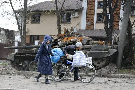 Local civilians walk past a tank destroyed during heavy fighting in an area controlled by Russian-backed separatist forces in Mariupol, Ukraine, Tuesday, April 19, 2022. Taking Mariupol would deprive Ukraine of a vital port and complete a land bridge between Russia and the Crimean Peninsula, seized from Ukraine from 2014. (AP Photo/Alexei Alexandrov)
