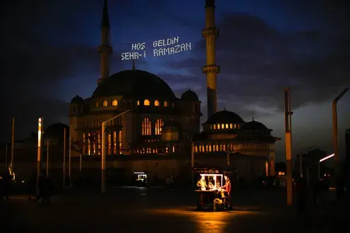 People buy roasted chestnuts next to Taksim mosque as the sun sets during the first day of the Muslim holy fasting month of Ramadan, in Istanbul, Turkey, Saturday, April 2, 2022. Lights on top read in Turkish: "Welcome Sehr-i Ramadan". (AP Photo/Francisco Seco)