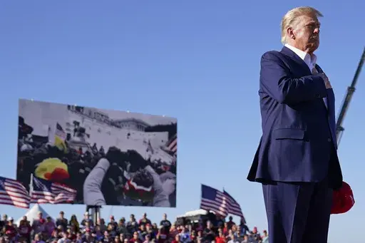 As footage from the Jan. 6, 2021, insurrection at the U.S. Capitol is displayed in the background, former President Donald Trump stands while a song, "Justice for All," is played during a campaign rally at Waco Regional Airport, Saturday, March 25, 2023, in Waco, Texas. The tune, “Justice for All,” is the Star-Spangled Banner and it was sung by a group of defendants jailed over their alleged roles in the January 2021 insurrection. The national anthem is overlaid with Trump reciting the Pledg