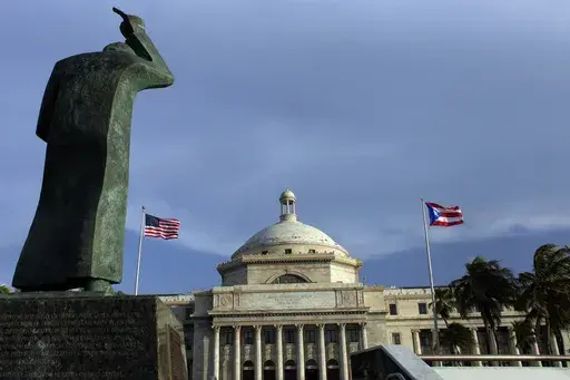 A bronze statue of San Juan Bautista stands in front the Capitol building flanked by U.S. and Puerto Rican flags, in San Juan, Puerto Rico. The Caribbean island's governor on Wednesday, July 24, 2024, signed a law that prohibits discrimination against people wearing Afros, curls, locs, twists, braids and other hairstyles in the racially diverse U.S. territory. (AP Photo/Ricardo Arduengo, File)