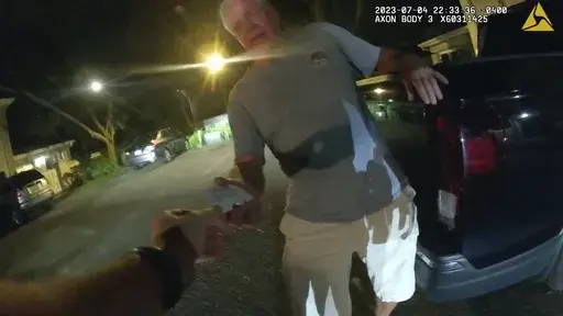 In this image from video provided by the Tampa Police Department, Joseph Ruddy, a prosecutor with the U.S. Attorney’s Office in Tampa, hands his business card to Officer Taylor Grant, outside his home in Temple Terrace, Fla., on the evening of July 4, 2023. When police arrived at his house to investigate a hit-and-run, Ruddy, one of the nation’s most prolific federal narcotics prosecutors, was so drunk he could barely stand up straight, leaning on the tailgate of his pickup to keep his balan